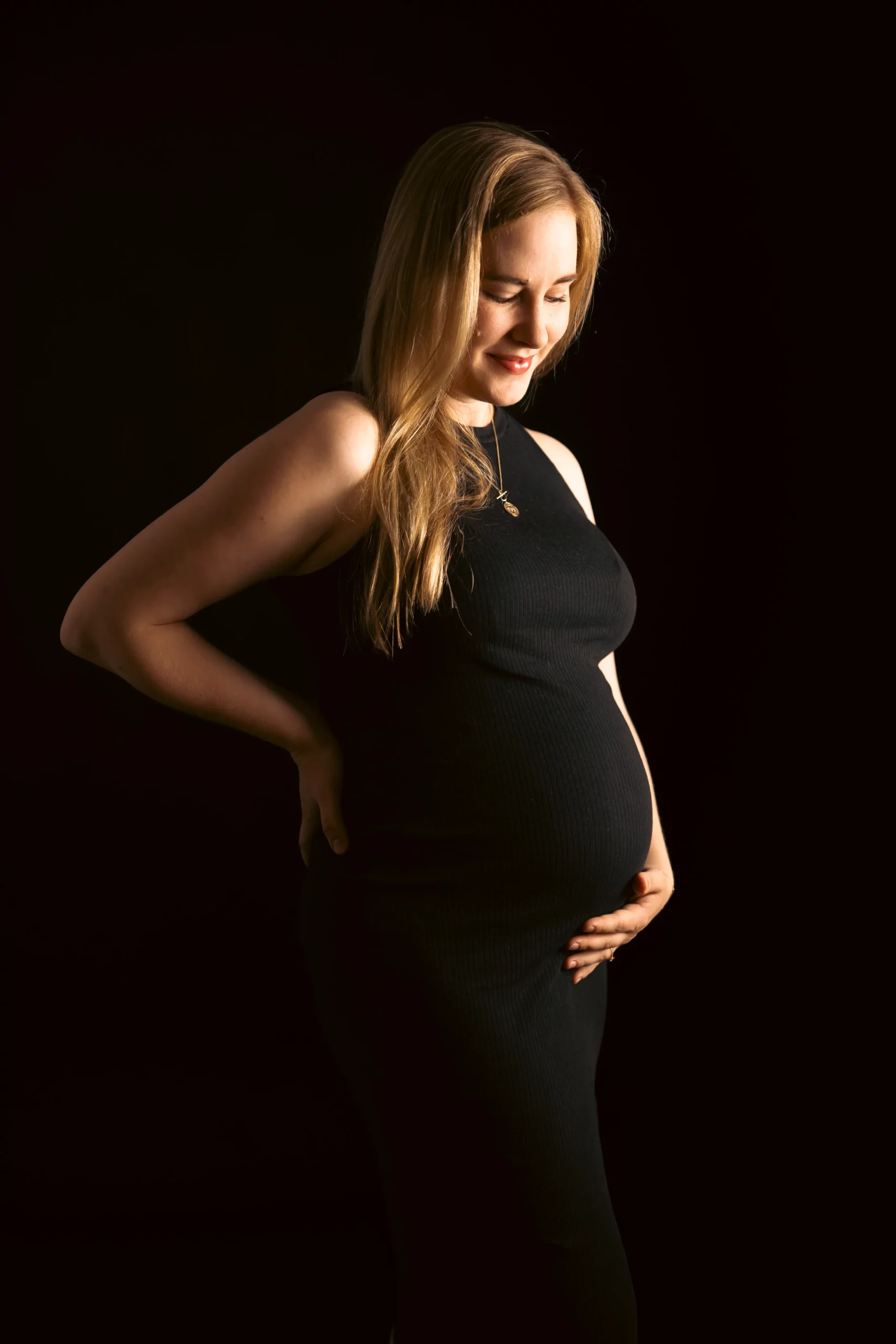Frau kurz vor der Geburt ihres Kindes beim Studio-Fotoshooting im schwarzem Kleid vor einem schwarzen Hintergund am lächeln.